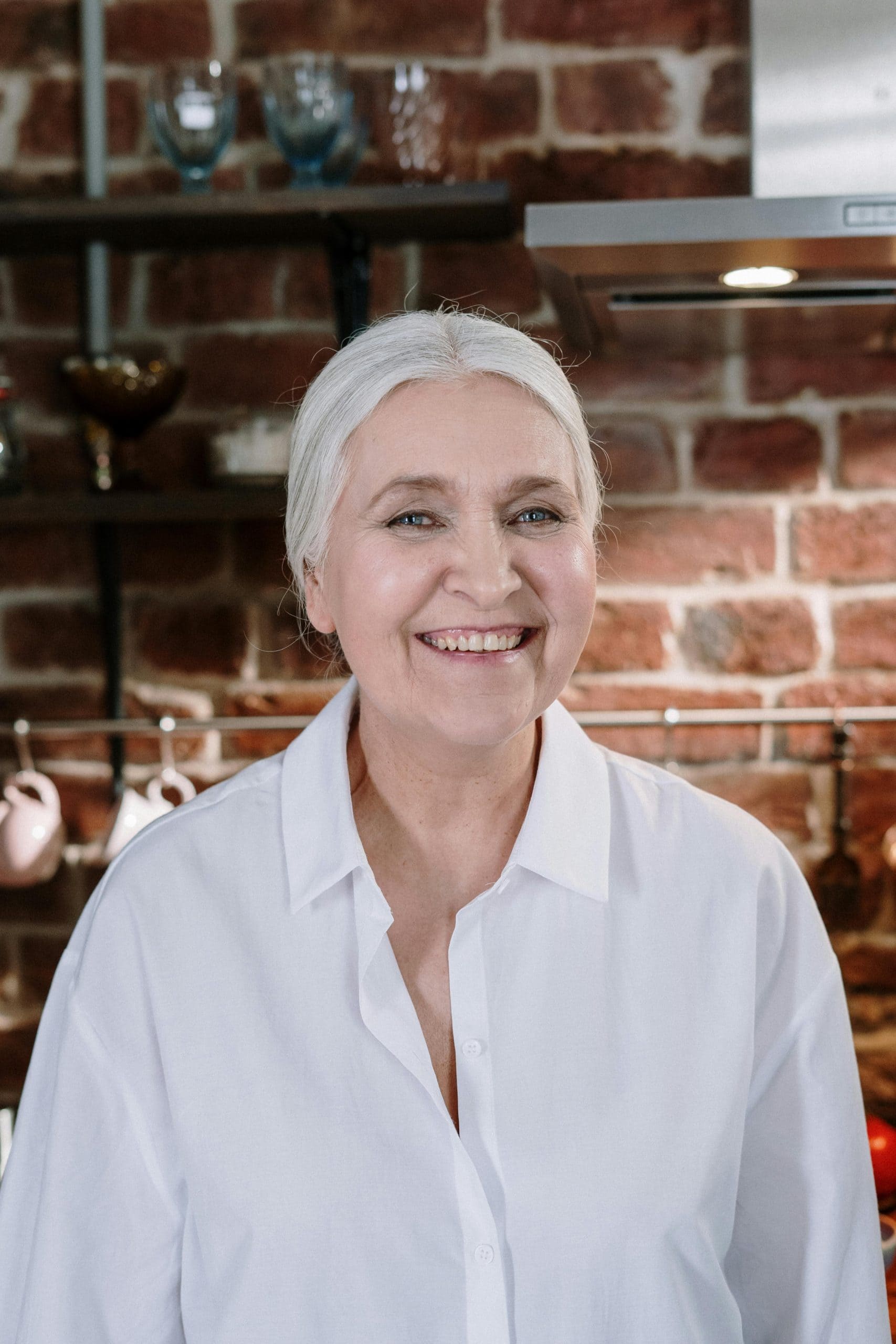 Senior woman smiling warmly in a cozy brick kitchen with rustic decor.